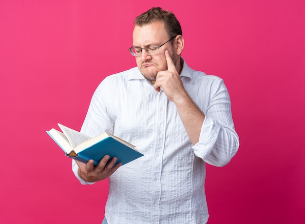 Man in white shirt wearing glasses holding book looking at it with pensive expression thinking standing on pink