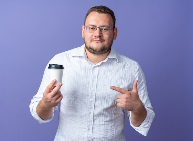 Uomo in camicia bianca con gli occhiali che tiene una tazza di caffè che punta con il dito indice sorridendo fiducioso in piedi sul muro blu