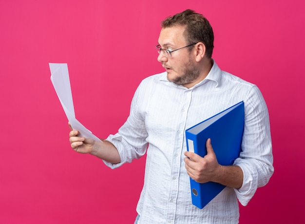 Man in white shirt wearing glasses holding office folder and documents looking at them amazed and surprised standing on pink