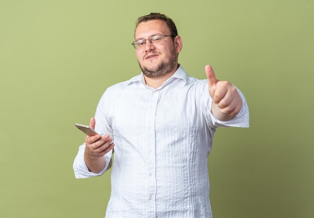 Man in white shirt wearing glasses holding smartphone looking at front smiling cheerfully showing thumbs up standing over green wall