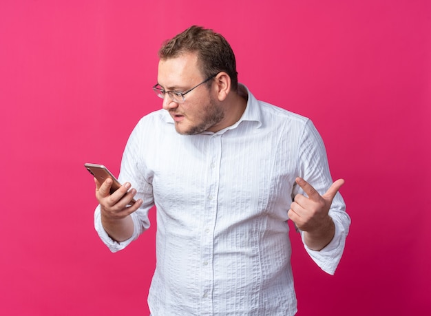 Uomo in camicia bianca con gli occhiali che tiene in mano uno smartphone guardandolo arrabbiato e frustrato in piedi sul muro rosa