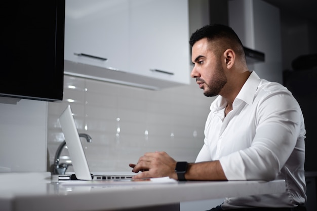 Man in white shirt websurfing with laptop in home kitchen