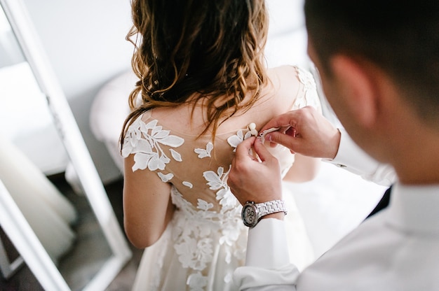 A man in a white shirt with a tie and a watch fastens buttons on the corset on the dress. Bride in wedding dress with lace standing at home.