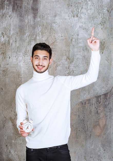 Man in white sweater holding an alarm clock and raising hand up.