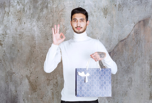 Man in white sweater holding a blue shopping bag and showing positive hand sign.