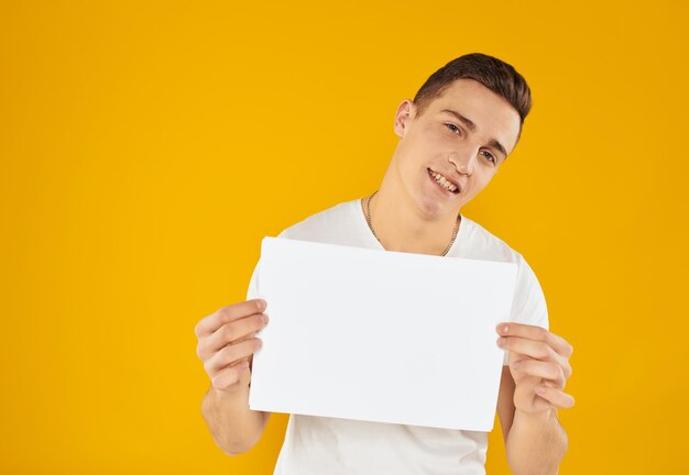 A man in a white t-shirt with a sheet of paper on a yellow background copy space mockup person