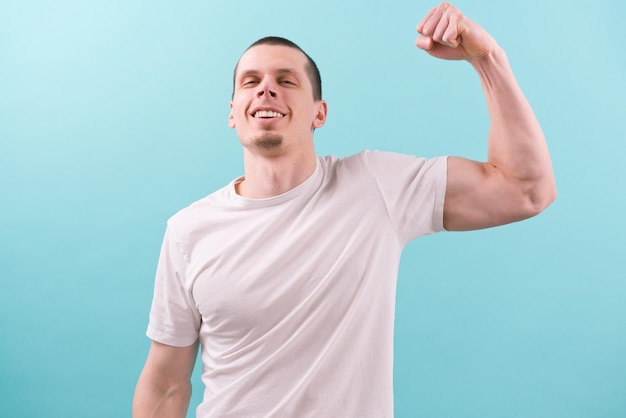 A man in a white tshirt smiles and shows his biceps on a blue background Confident Emotional Facial Hands Human Look Posing Power Sexual Shoulder Portrait