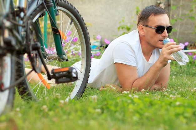 Man in white tshirt and sunglasses with bottle of water lying on green grass with bicycle