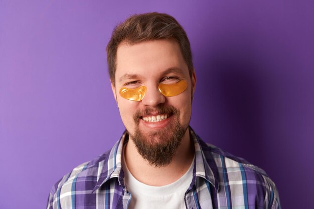 Man in white tshirt with eye patches stand against violet background. Beauty treatment spa wellbeing concept. Self care routine