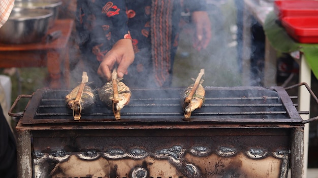 A man who is grilling fish traditionally uses a grill to sell\
and see the smoke. sea food.