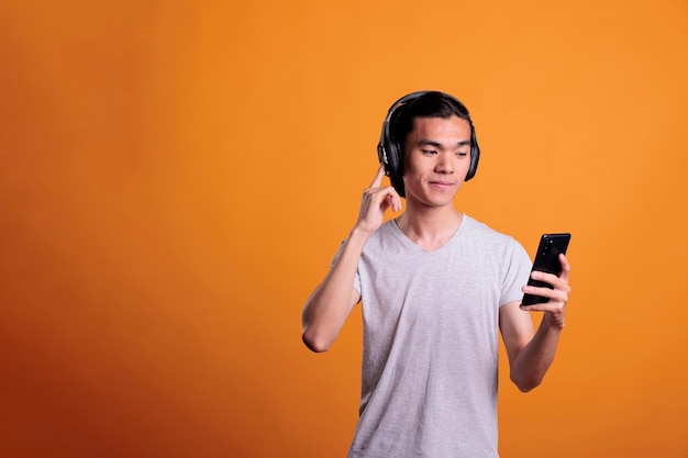 Man in wireless headphones listening to podcast, smiling asian\
teenager enjoying music. young person standing in earphones,\
holding smartphone, watching video on mobile phone