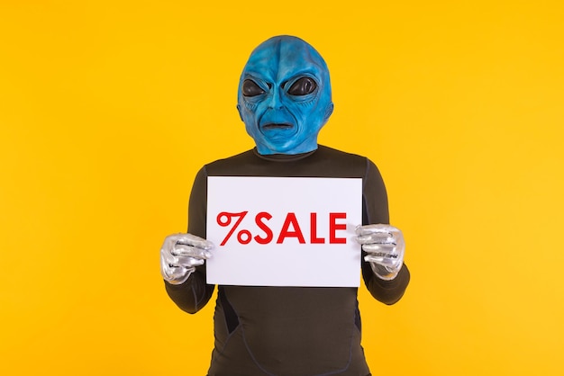 Man with an alien mask with a blue head holding a white sign with red letters that reads 39 Sale39 Concept of bizarre extraterrestrial funny informative weird and weird