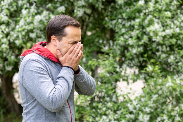 A man with an allergy or cold sneezes against a flowering tree