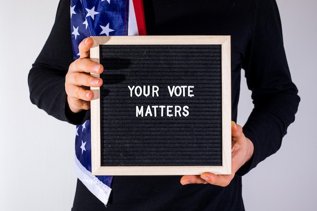 Foto uomo con bandiera americana in possesso di una lavagna con testo your vote matters su sfondo bianco