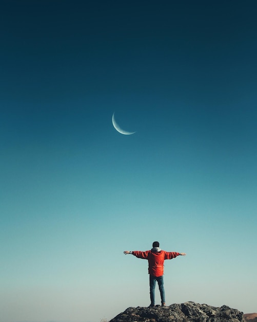 man with arms outstretched standing on rock against clear blue sky