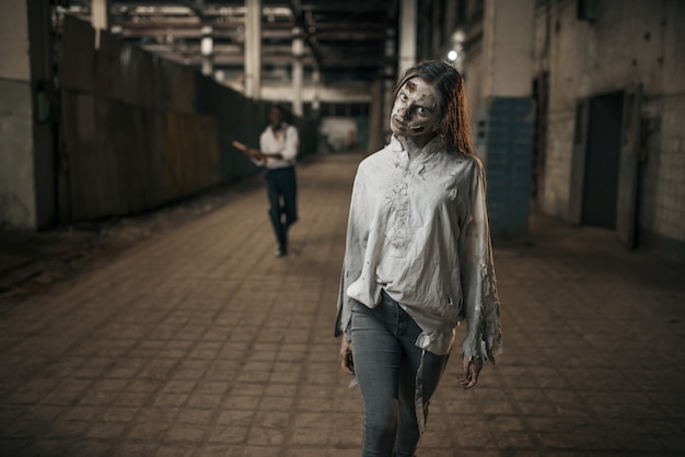 Man with axe catching up female zombie in abandoned factory, scary place. Horror in city, creepy crawlies, doomsday apocalypse, bloody evil monsters