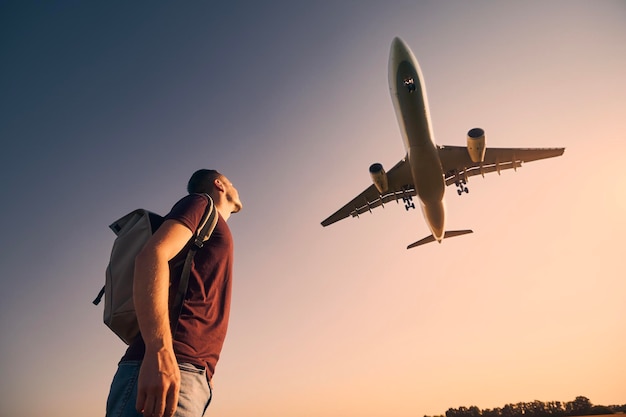 man with backpack looking up to airplane landing at airport during beautiful summer sunset