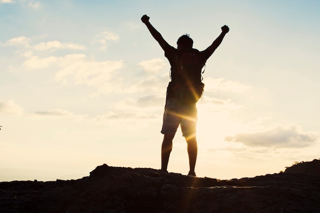 Man with backpack putting his hands up  and standing on cliff at sunset time