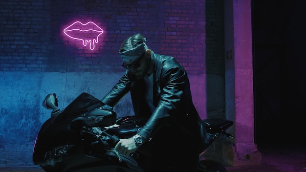 A man with a bandama sits on a sports motorcycle in sunglasses against a brick wall with a neon sign