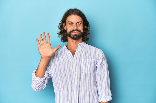 Man with beard in blue striped shirt blue studio smiling cheerful showing number five with fingers