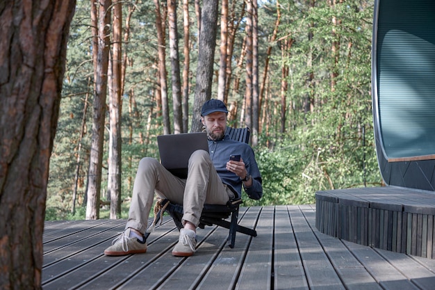 a man with a beard and in a cap sits in nature and works in a laptop