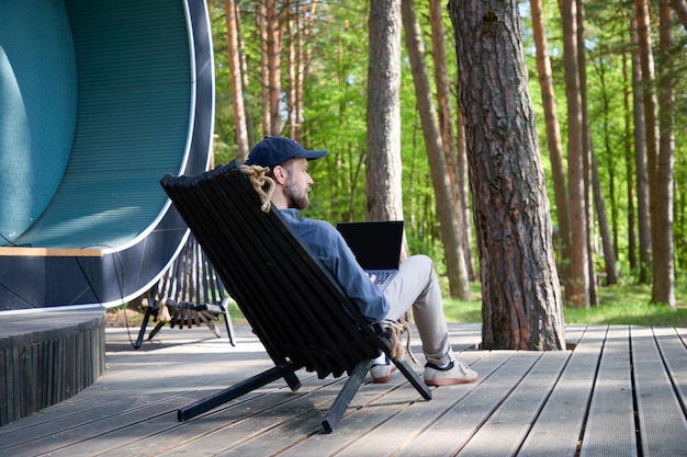 мужчина с бородой и в кепке сидит спиной на природе и работает за ноутбуком
