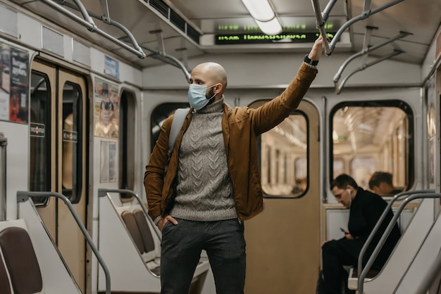 A man with a beard in a face mask to avoid the spread of coronavirus is holding onto the handrail in the center of a subway car. A guy in a surgical mask against COVID-19 is standing on a metro train.