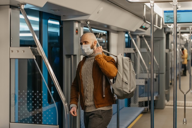 A man with a beard in a face mask to avoid the spread of coronavirus is putting on a gray backpack while riding a subway car. A bald guy in a surgical mask is keeping social distance on a train.