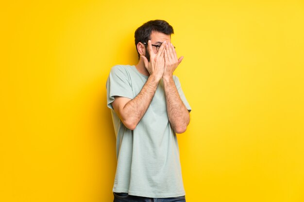 Man with beard and green shirt covering eyes by hands and looking through the fingers