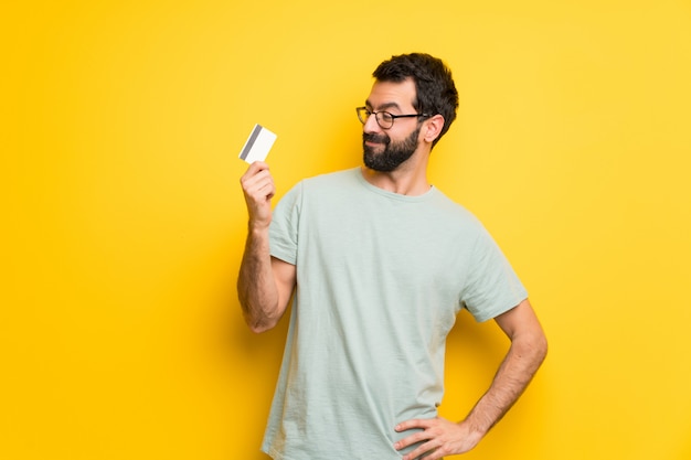 Man with beard and green shirt holding a credit card and thinking