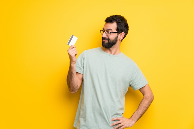 Uomo con la barba e camicia verde in possesso di una carta di credito e il pensiero