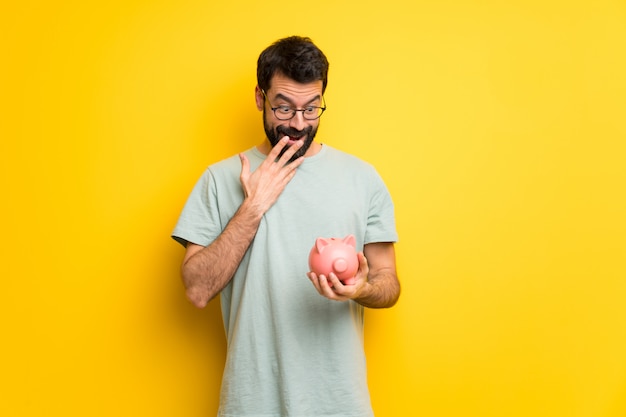 Man with beard and green shirt surprised while holding a piggybank