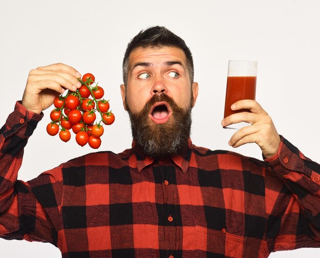 Man with beard holds glass of juice and vegetables isolated on white background Guy holds homegrown harvest Farming and autumn concept Farmer with surprised face shows bunch of red cherry tomatoes