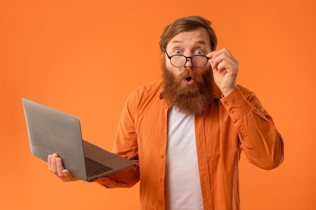 Man with beard holds laptop looks with shocked expression studio