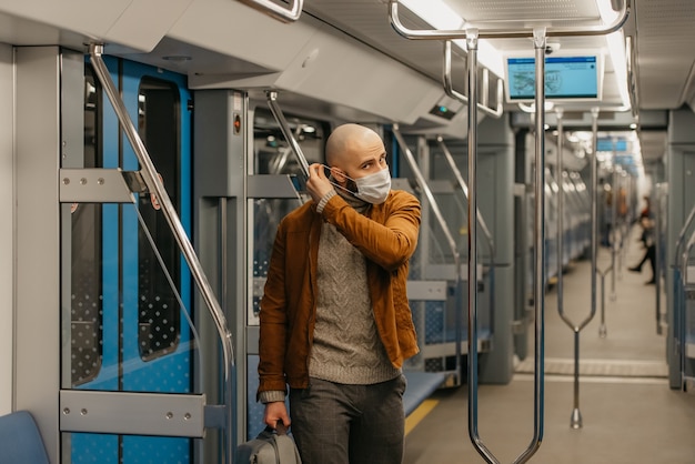 A man with a beard is putting on a medical face mask to avoid the spread of coronavirus in a subway car. A bald guy in a surgical mask against COVID-19 is keeping social distance on a train.