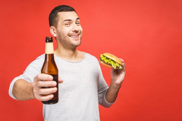 Man with a beard isolated over red background holds a hamburger and a bottle of beer, portrait