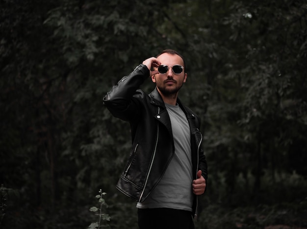 A man with a beard and a leather jacket straightens his glasses on the background of the forest