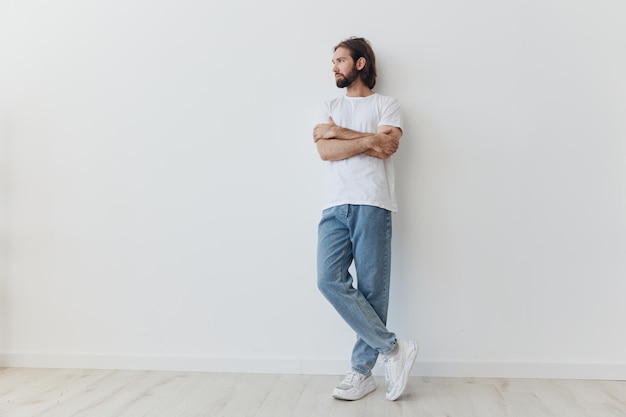 Photo a man with a beard and long hair in a white tshirt and blue jeans stands against a white wall leaning fulllength on it relaxed style