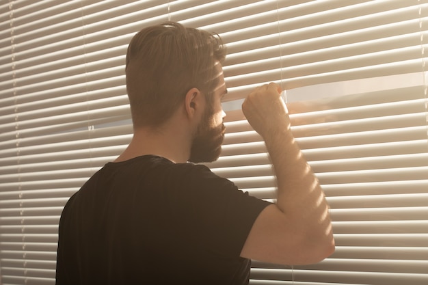 Photo man with beard peeks through hole in the window blinds