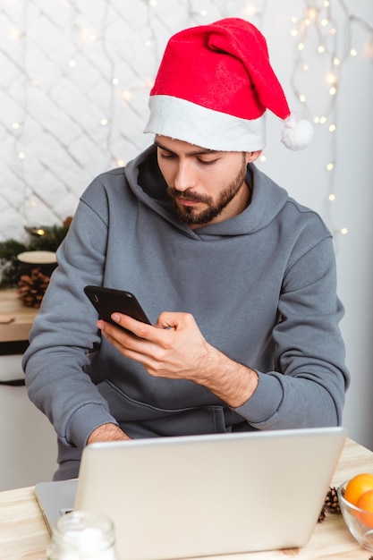 Man with beard sitting kitchen use laptop buy gifts Internet hold mobile phone garlands atmosphere