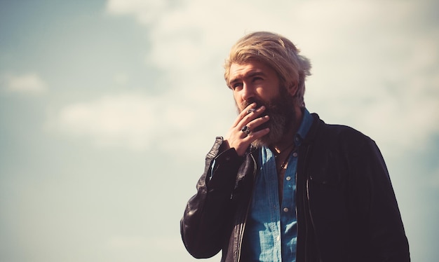 수염 흡연 담배를 가진 남자 자연 배경에 흐린 하늘에서 재킷에 젊은 수염된 hipster