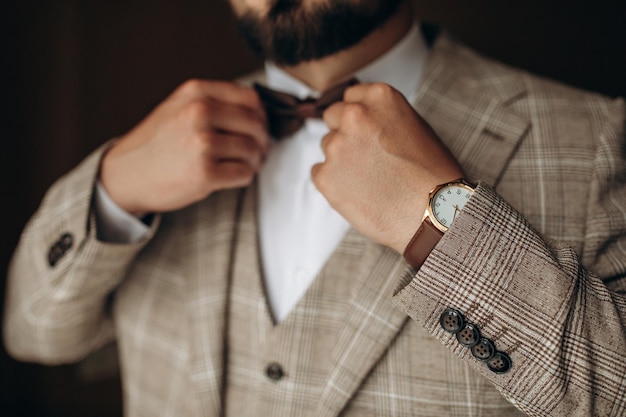 A man with a beard in a stylish suit with a watch on his arm fixes a bow tie Morning of the groom Close up detail of man fashion