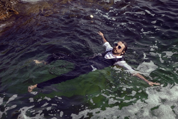 Photo man with a beard and sunglasses in clothes a vest and a white shirt swims in sea among the rocks in summer