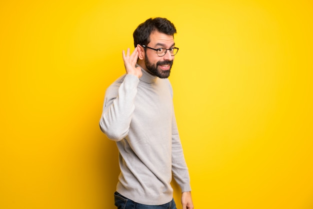 Man with beard and turtleneck listening to something by putting hand on the ear