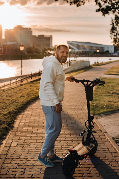 A man with a beard in white clothes rides around the city on an electric scooter at sunset