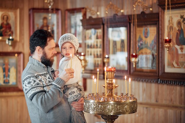 A man with a beard with a little girl in his arms stands before the icons and prays in the Orthodox Church