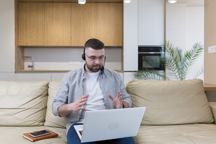 Photo a man with a beard works from home sitting on the couch uses a laptop and a headset with a microphone conducts an online consultation