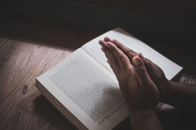 Man with Bible praying, hands clasped together on her Bible on wooden table.