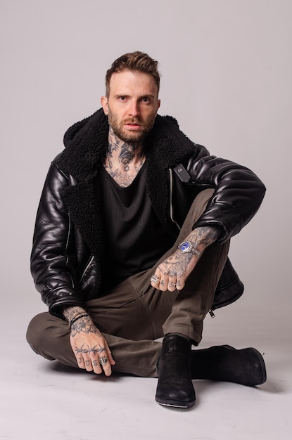a man with a black leather jacket sits on the floor with a tattoo on his arm.