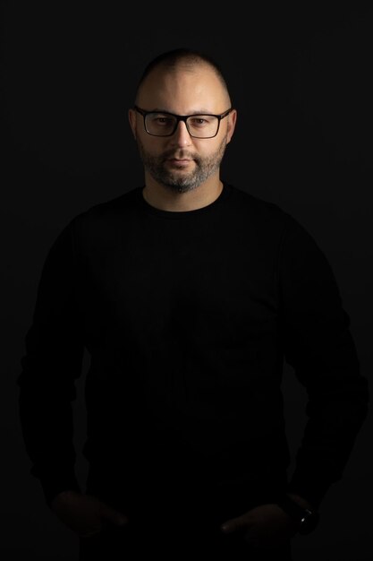 A man with black-rimmed glasses and a black sweater keeps his hands in his pockets